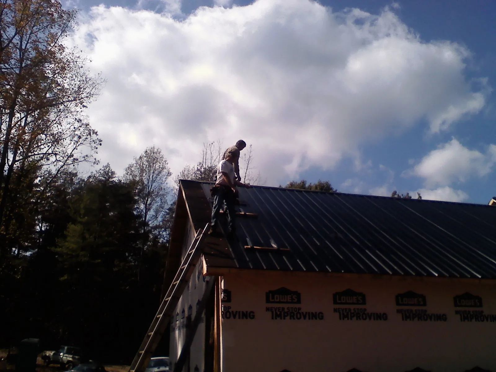 Roofer fastening dark metal panels high against a cloudy sky, from a Roof Inspection job in Old Forge
